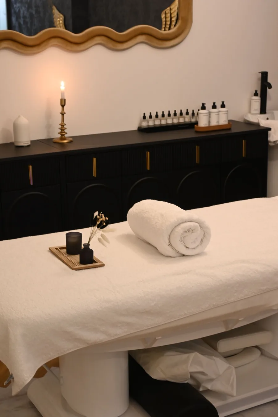 A treatment table with a rolled towel, a candle and small oil bottles on a dark cabinet