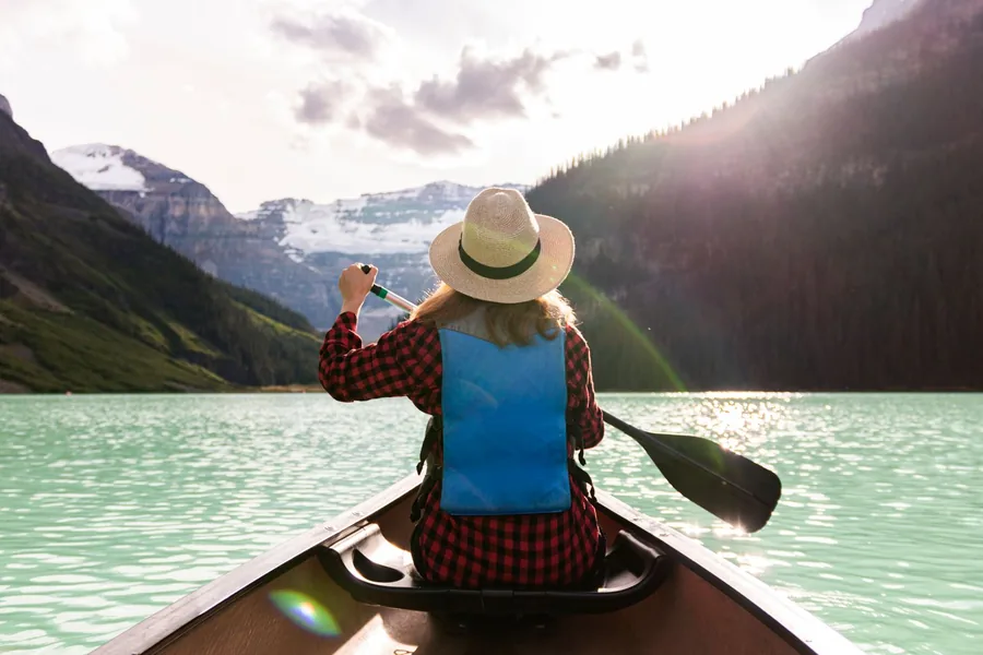 Seen from the back of a canoe, a paddler in a straw hat crosses turquoise Lake Louise toward snowy mountains